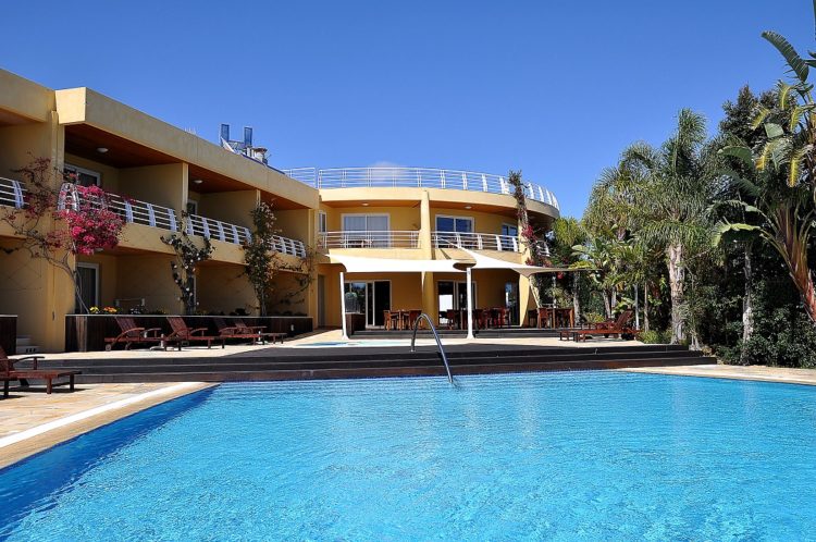 Blue skies and a large blue swimming pool dominate the scene with a backdrop against the aparthotel