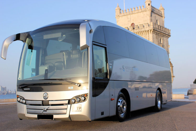 A large silver coach shining in the afternoon sun with the backdrop of a castle and the blue sea